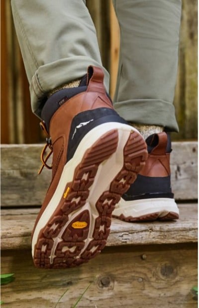 Brown leather hiking boots with rugged soles, paired with rolled light green pants, stepping on wooden outdoor stairs.