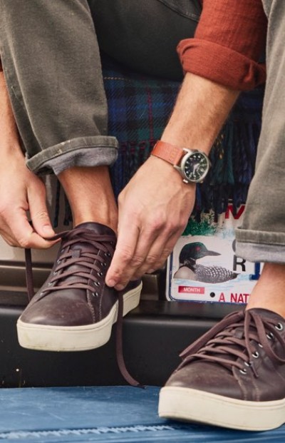 Person tying brown sneakers while sitting on a bumper with a loon-themed license plate; wearing a watch on their wrist.