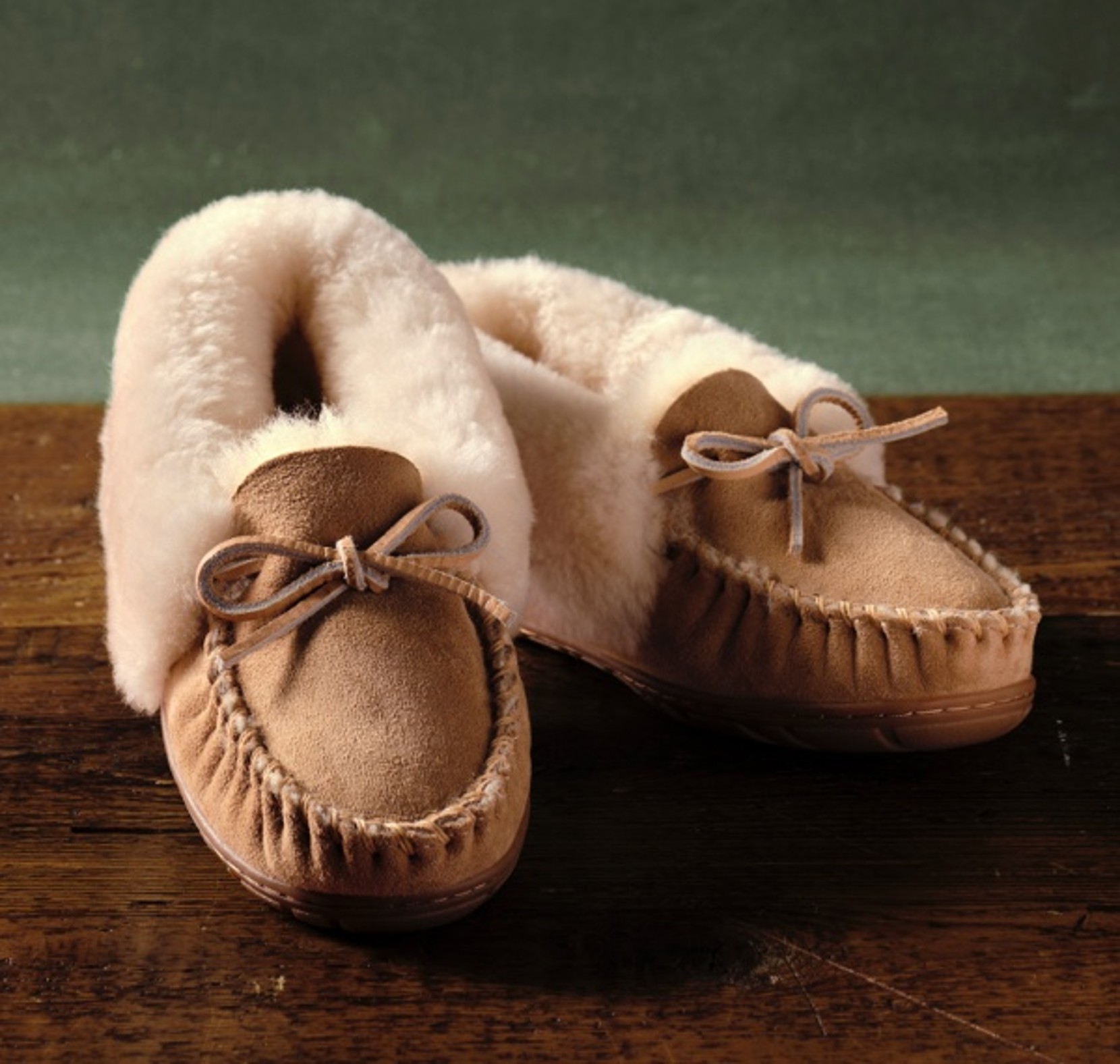 Brown suede moccasin slippers with fleece lining and rubber soles on wood.