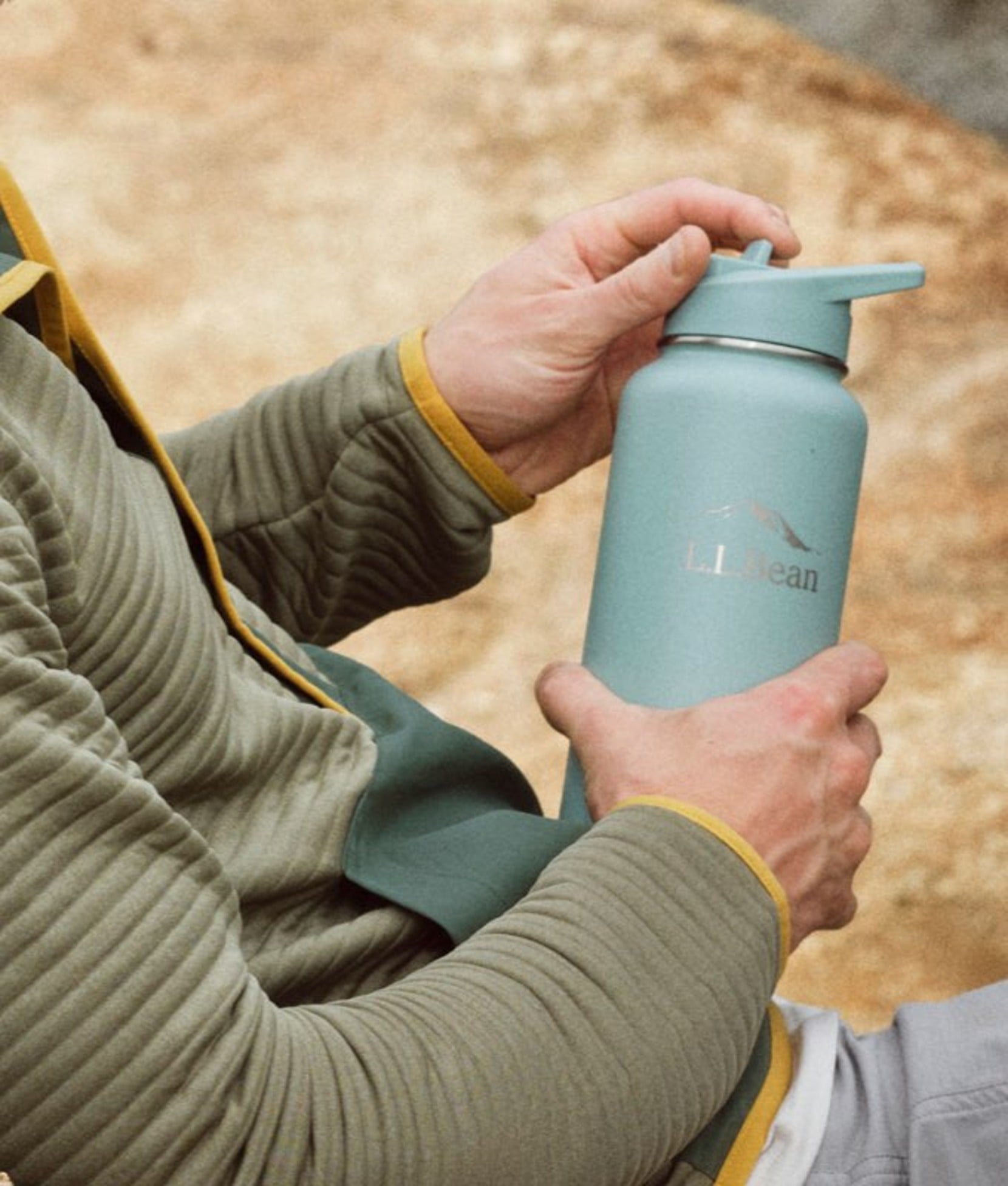 Person in green airlight top holding blue water bottle outdoors near sandy ground.