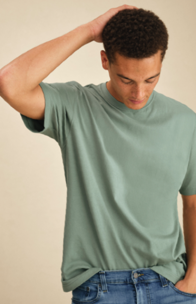 A man wearing a sage green short-sleeve T-shirt paired with blue jeans, styled in a casual look against a neutral background.
