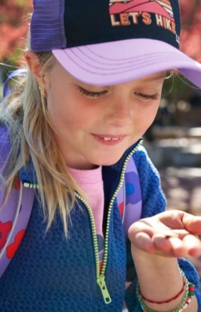 Child wearing a navy textured jacket, a purple backpack, and a cap that reads “LET’S HIKE,” holding something in hand.