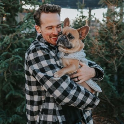 Man holding a bull dog outside among fir trees.