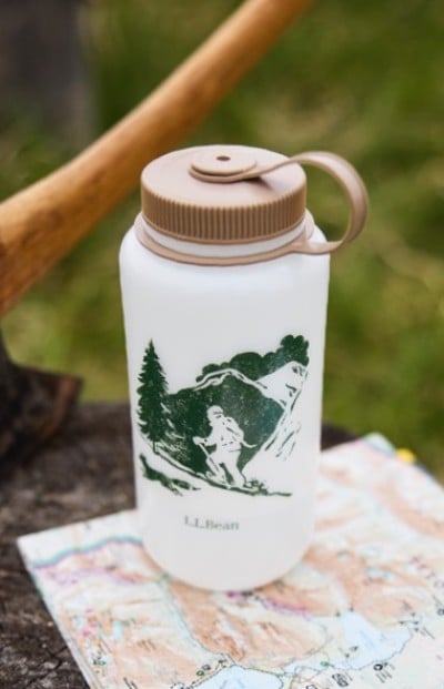 White L. L. Bean water bottle with green hiker graphic sits on a colorful map, with an axe handle in the background.