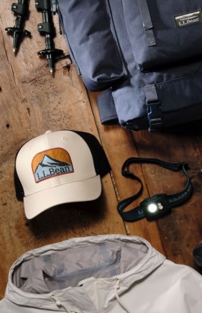 Outdoor gear laid out on a wooden surface, including a backpack, cap, headlamp, trekking poles, and a light jacket.
