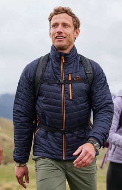 A man in blue quilted jacket and green pants hikes with a backpack under a cloudy sky, surrounded by grassy hills.