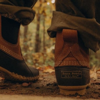 Close-up of person wearing brown and black Bean Boots, standing on a forest floor covered in fallen leaves.