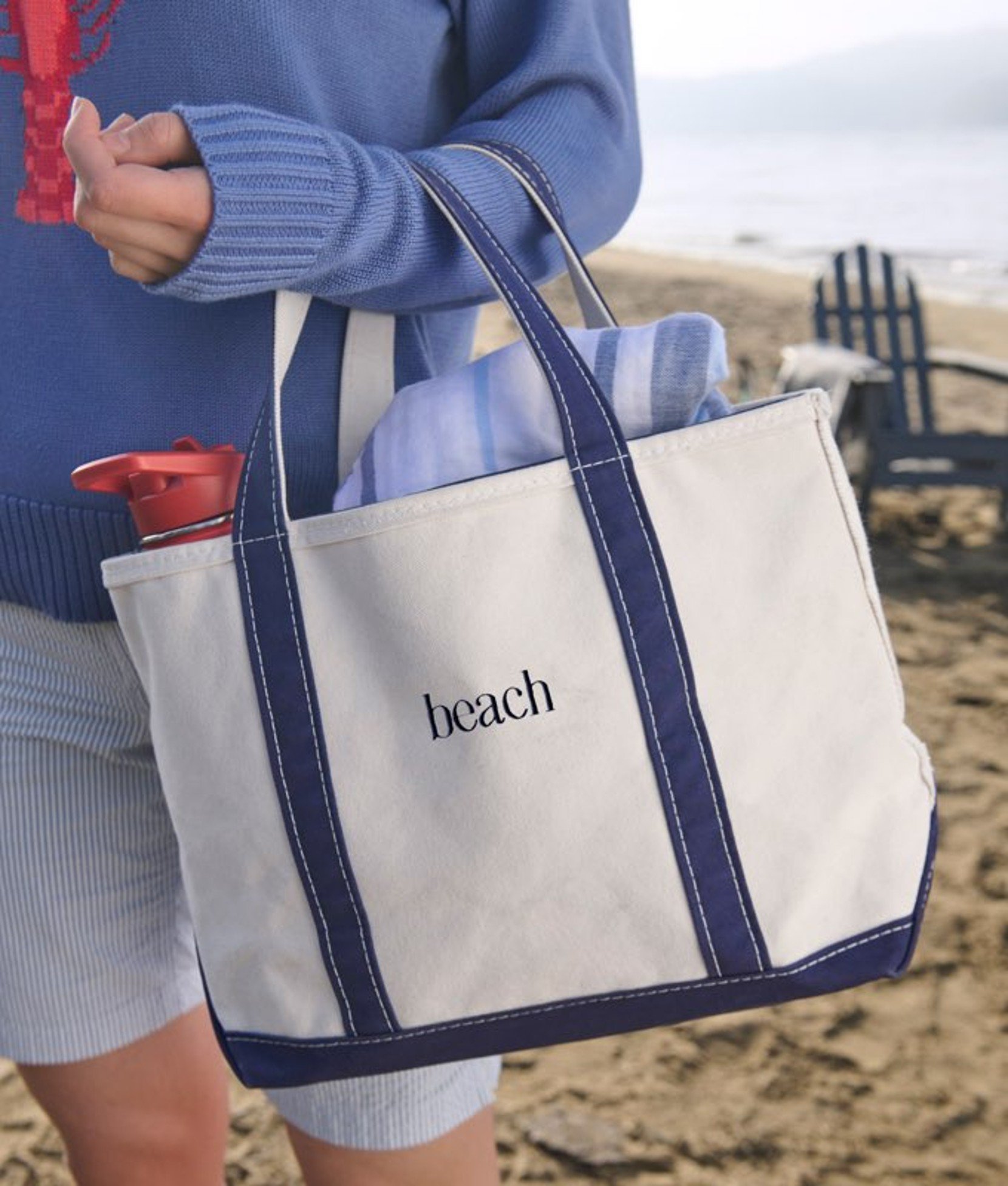 Canvas tote bag with sturdy handles resting on sand near the beach, with water blurred in the background.