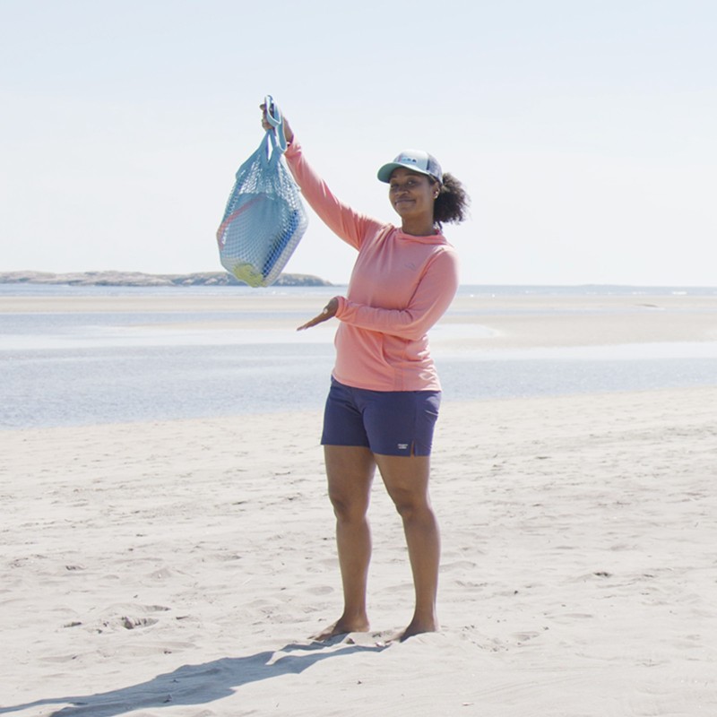 Stephanie standing on the beach showing a mesh bag filled with beach toys.