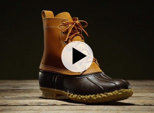 Brown and tan leather Bean Boot with yellow laces on a wooden surface, with a play button overlay.