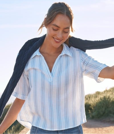 Waist-up shot of woman on a beach path, holding the sleeves of a lightweight sweater draped on her shoulders.