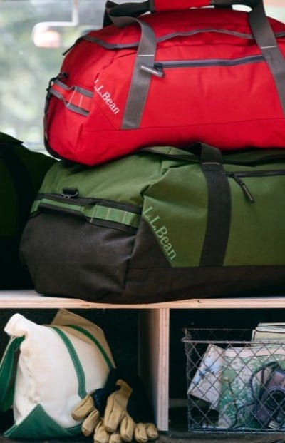 Two L.L.Bean duffel bags, one red and one green, are stacked on a shelf with gloves, a canvas bag, and a wire basket below.