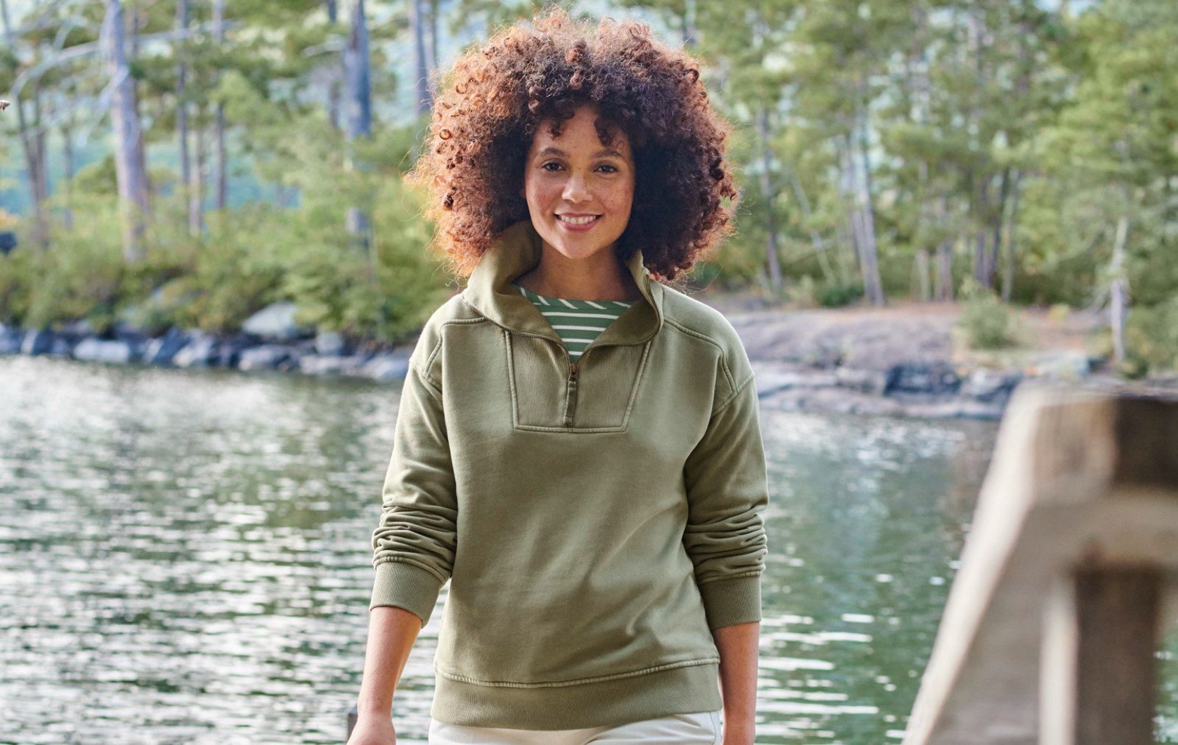 Person walking beside a lakeside dock, wearing an olive pullover and striped shirt in a wooded setting.