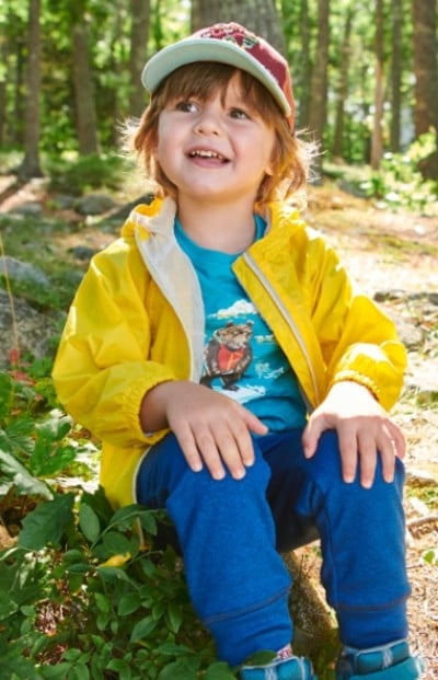 Toddler sitting in the woods wearing a yellow jacket, blue pants and a hat.