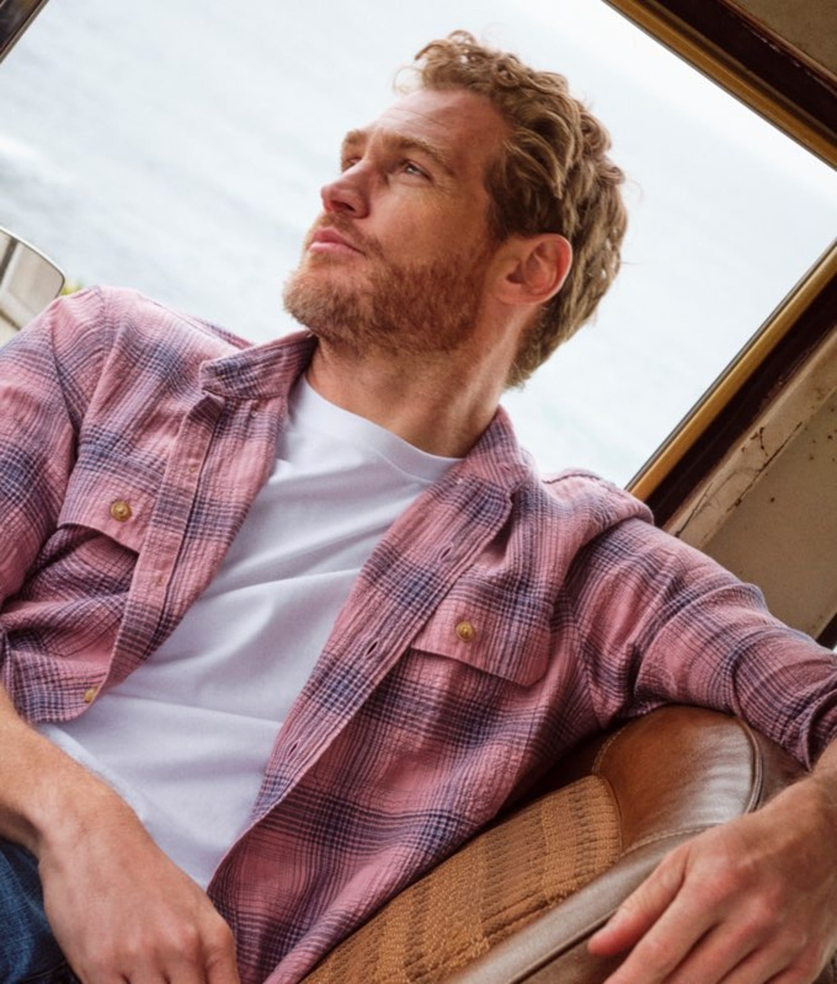 A handsome gent in mauve plaid shirt leaning back to a window showing ocean blue water.