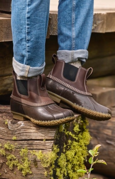 A person in brown leather Chelsea L. L. Bean boots and rolled-up blue jeans stands on a wooden surface with moss and plants below.