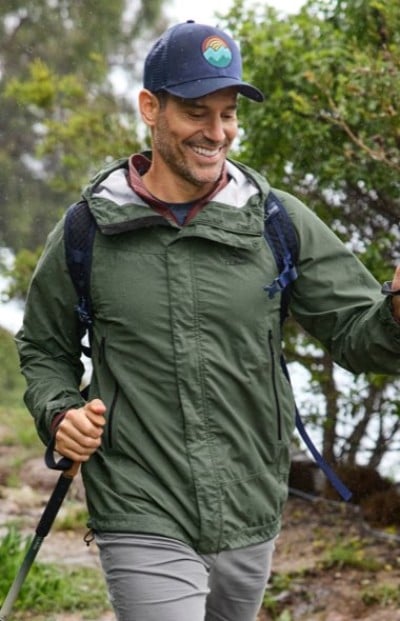 Person hiking on a trail in a green jacket and cap, carrying a backpack and using a trekking pole.