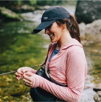A smiling woman standing in a river with a fly fishing rod under one arm tying a fly on her line.