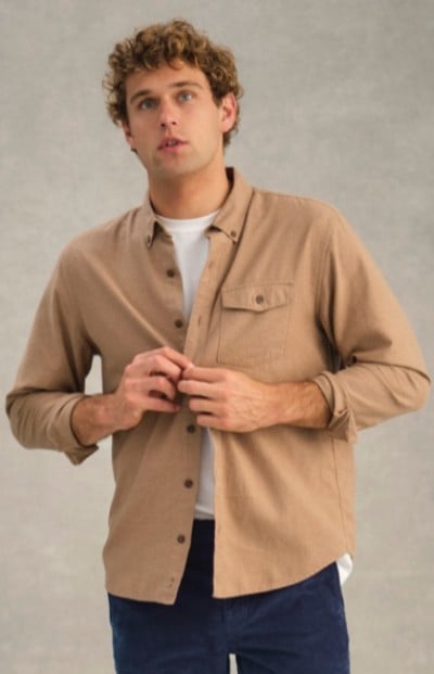 Person in beige button-up shirt over white t-shirt and dark blue pants stands against a plain gray background.