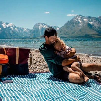 Man hugging a small girl sitting on a picnic blanket, beautiful snow capped mountains and water in the background.