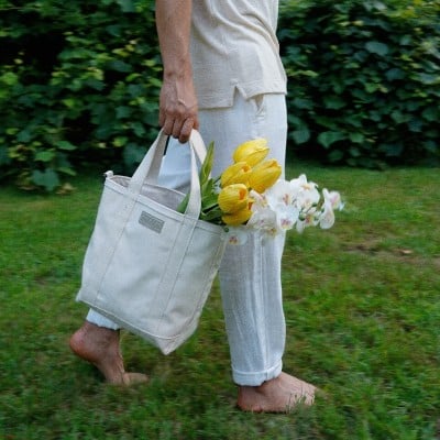 A waist-down shot of a person walking barefoot through the grass carrying a canvas tote bag with yellow tulips and white daffodils.