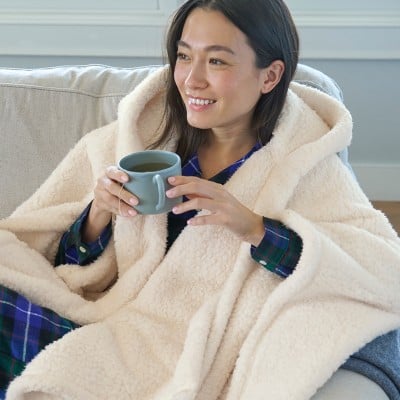 A woman wrapped in a cozy throw sits and smiles and she holds a coffee mug.