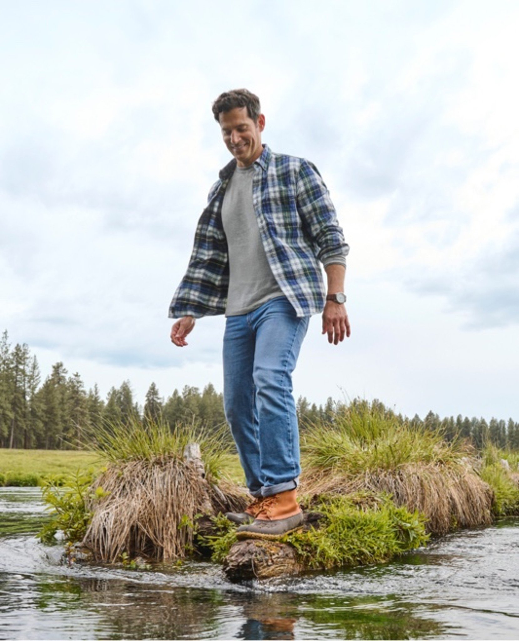 Person balances on grassy mound in plaid shirt and boots.