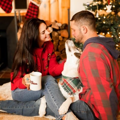 A couple sitting on the floor in front of a holiday tree petting a dog.