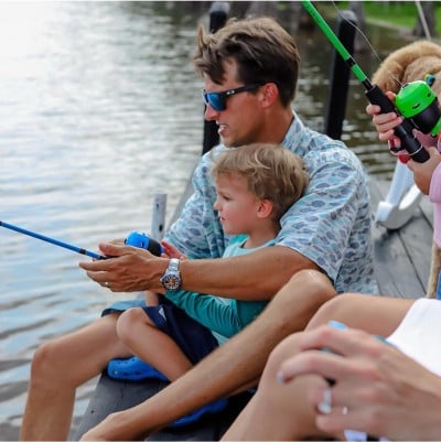 A man sitting on a dock helping a young boy in his lap to fish.