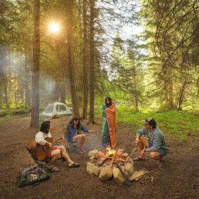 A group of friends around a campfire, one playing a guitar, one wrapped in a blanket, one pokes at the fire.