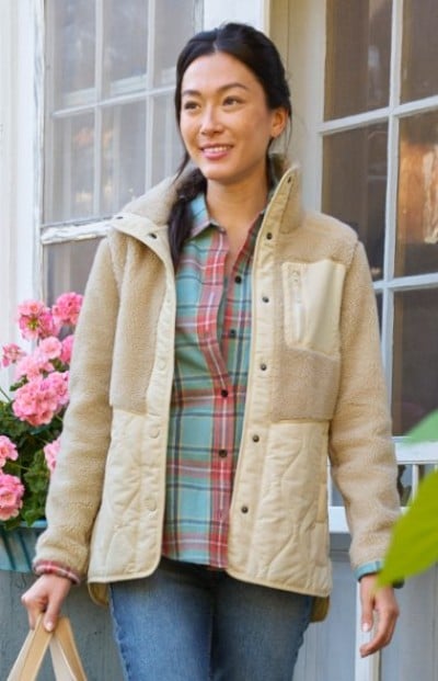 Woman wearing a beige fleece jacket and plaid shirt stands outdoors near pink flowers.