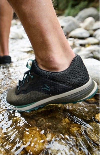 A person wearing black L L Bean sneakers steps into a shallow stream with rocks and water visible.