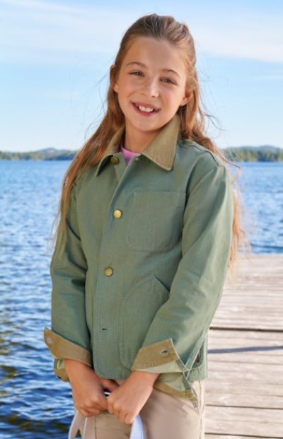 A child in a light green jacket with a tan collar stands on a dock near the water.