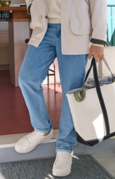 Person wearing light-wash jeans, white sneakers, and a beige jacket while holding a large canvas tote bag indoors.