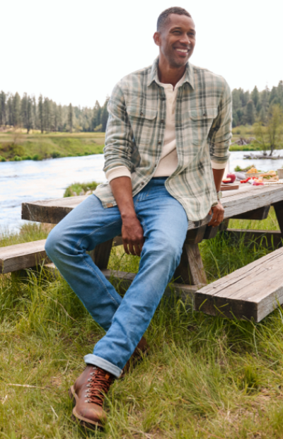 A man wearing blue jeans, a plaid shirt, and leather boots sits on a picnic table near a river.