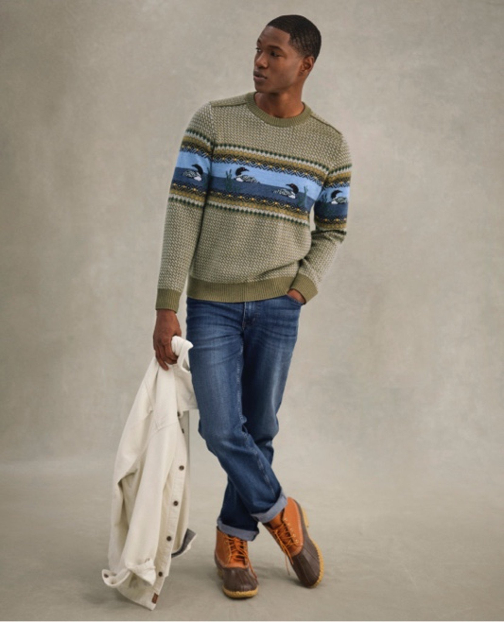 Person holds white jacket, wears green duck-patterned sweater and jeans.
