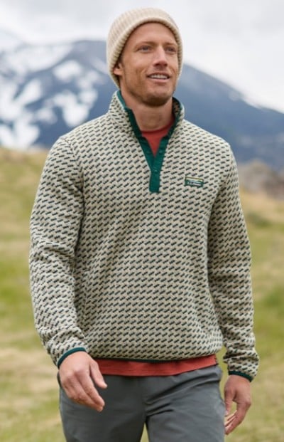 A man wearing a patterned sweater fleece, orange shirt, beige beanie and gray pants walks outdoors with mountains in the background.