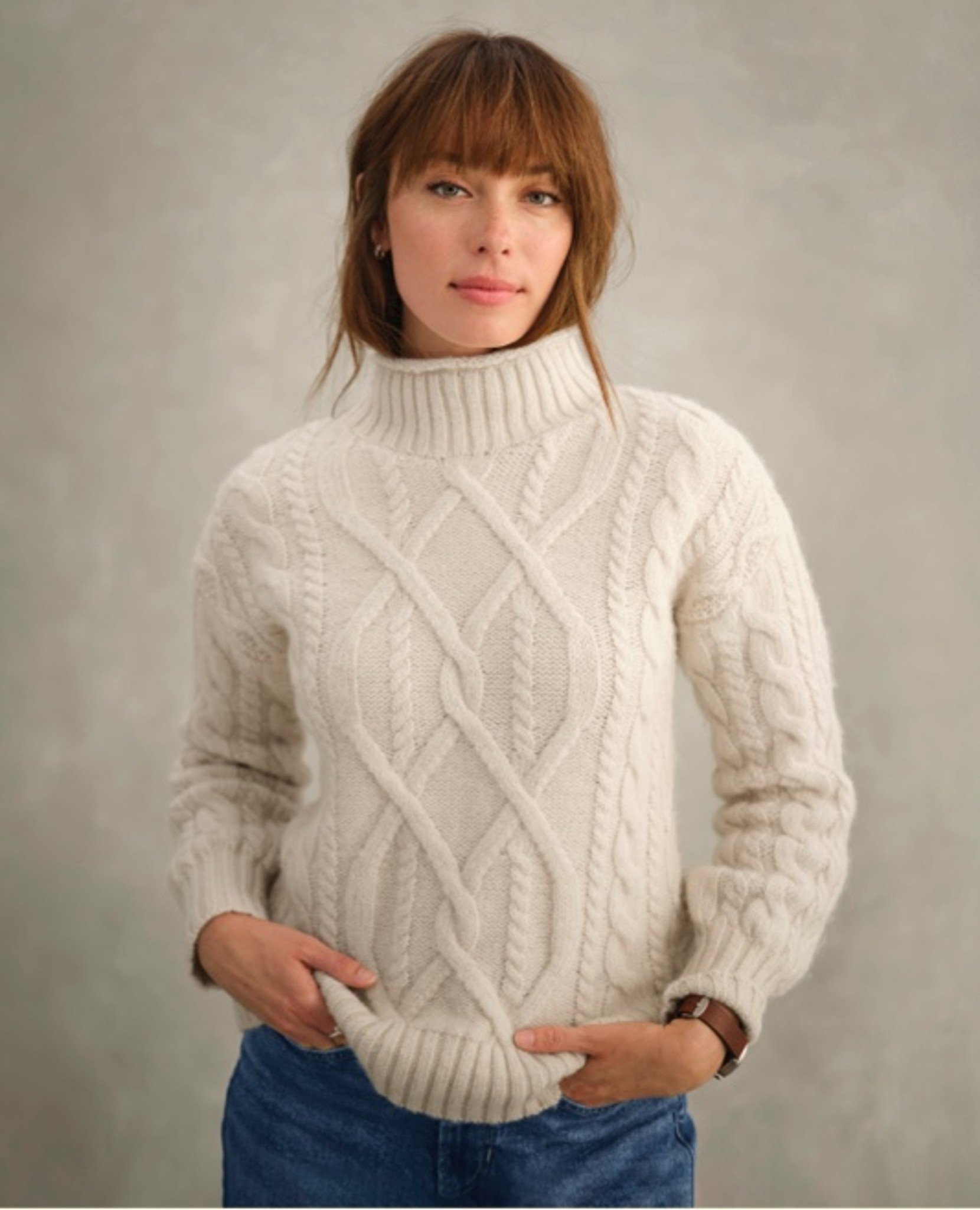 Person in cream cable-knit turtleneck and jeans looks at camera.