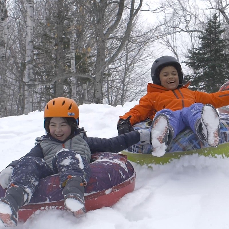 Children in a winter setting slide on snow using L L Bean Sonic Snow Tubes.