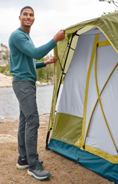 Person wearing a teal long-sleeve shirt and gray pants sets up a green and white tent near a lake in a forested area.