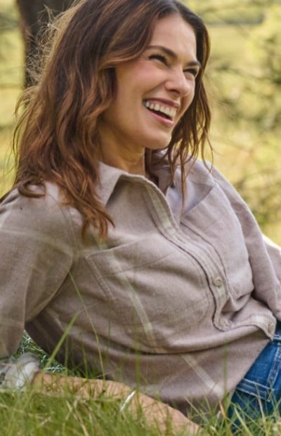 Woman with long hair in beige shirt and jeans sits on grass outdoors, smiling and looking to the side. 