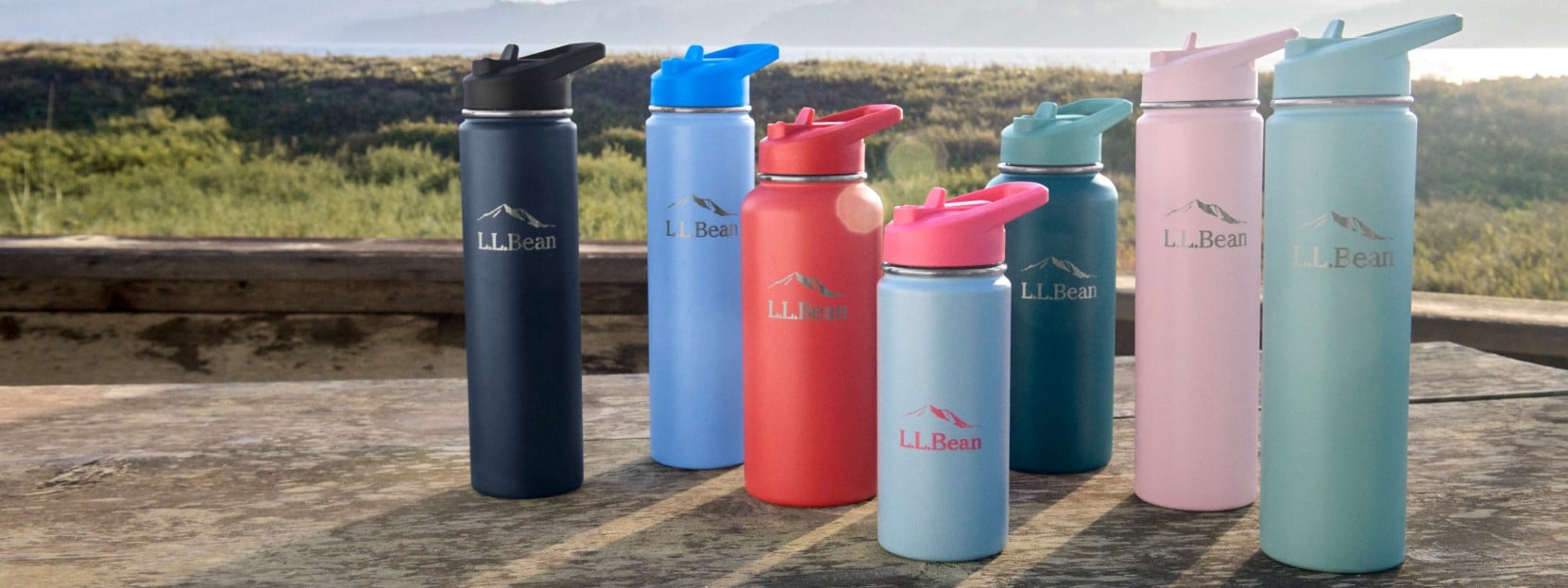 Seven colorful L L Bean water bottles with flip lids on a wooden table outdoors, greenery in sunlight behind. 