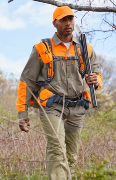 A person wearing an orange cap and jacket, carrying a rifle, walking through a grassy area.