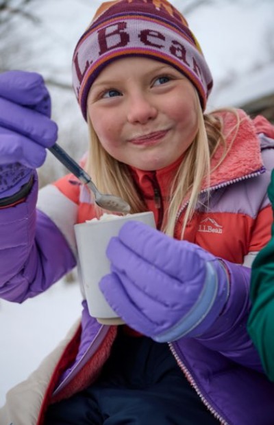 Child in winter clothing and hat holding a cup while wearing purple gloves in the snow.