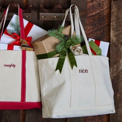 Two canvas tote bags labeled “naughty” and “nice,” filled with wrapped holiday gifts and greenery.