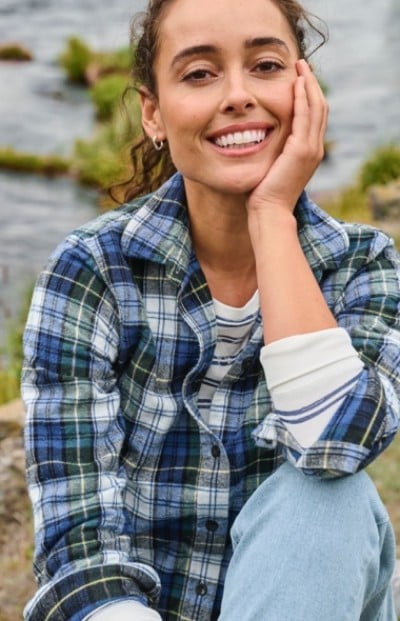 Woman in plaid shirt and jeans sits by a river, smiling with head resting on hand in a relaxed outdoor setting.