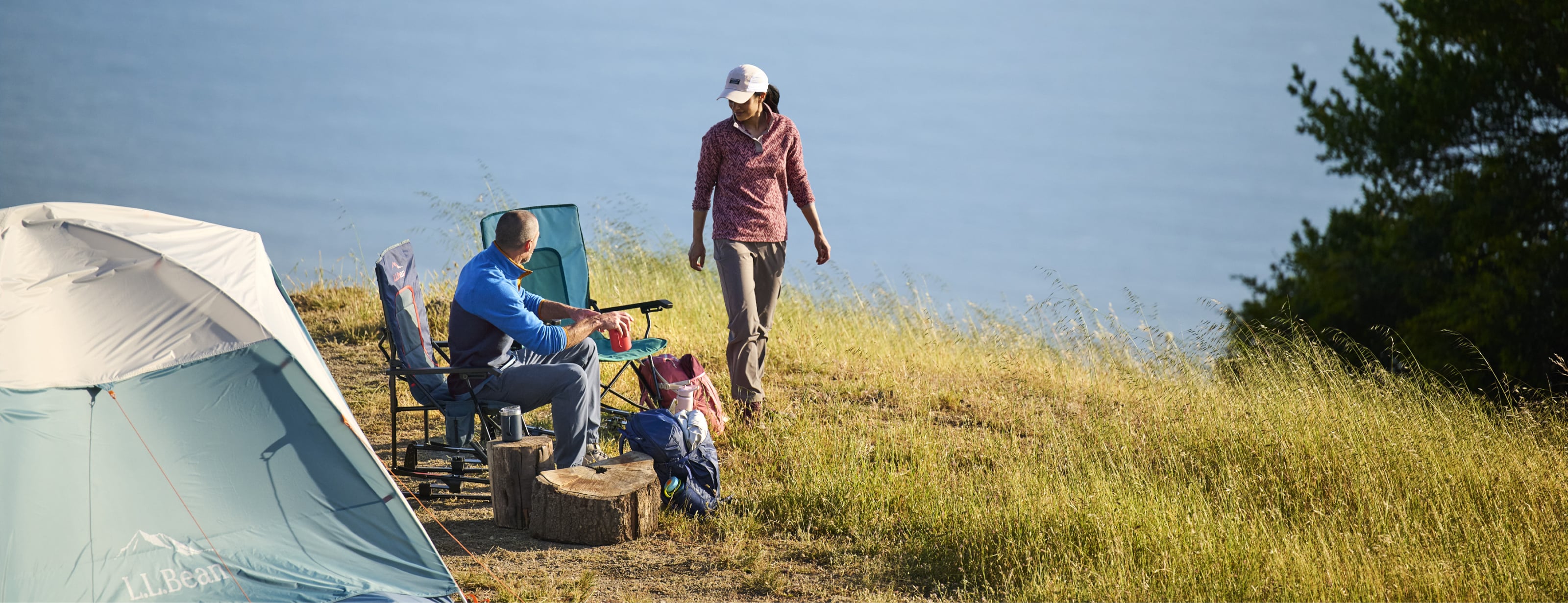 Family campsite with a tent, chairs and campers in an open field near water.