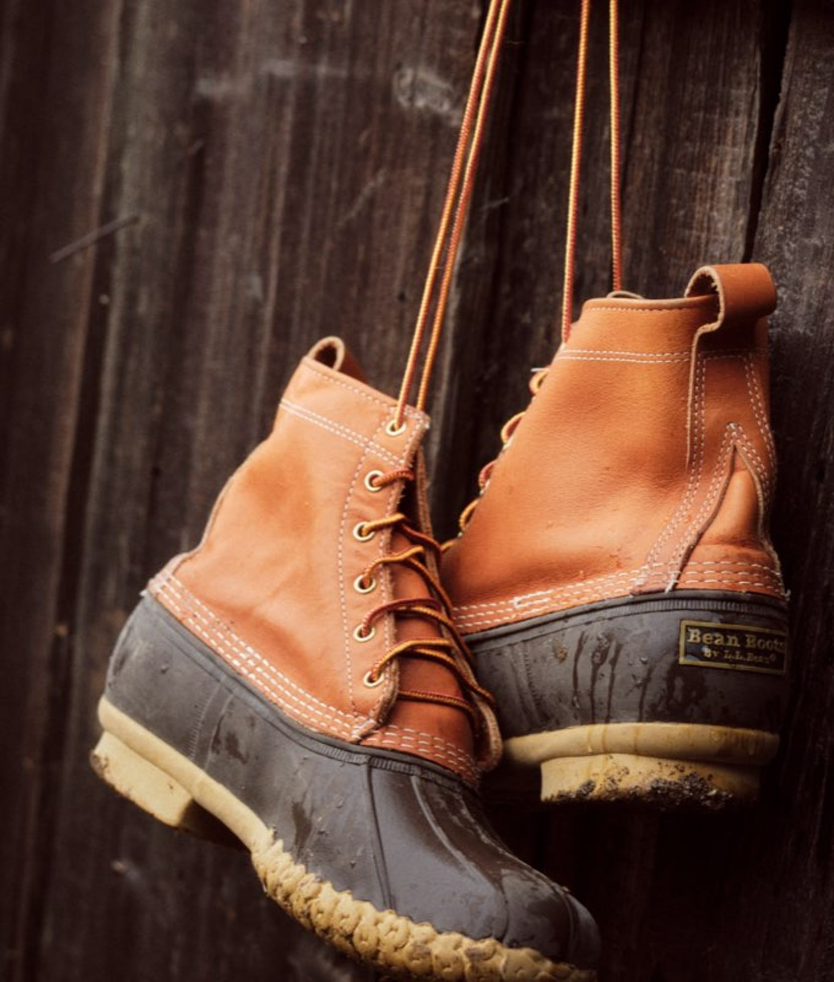A pair of brown leather L L Bean Boots hanging from their laces next to rustic wood wall.