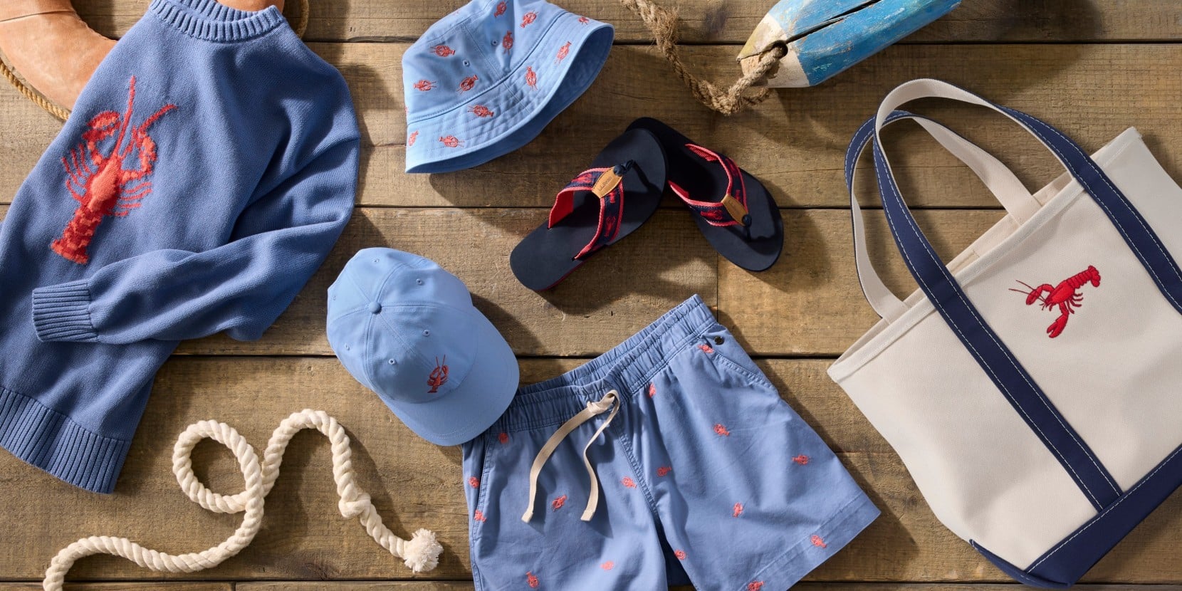 Flat lay of blue lobster-print sweater, shorts, hat, cap, flip-flops and tote with buoy and rope on wood.