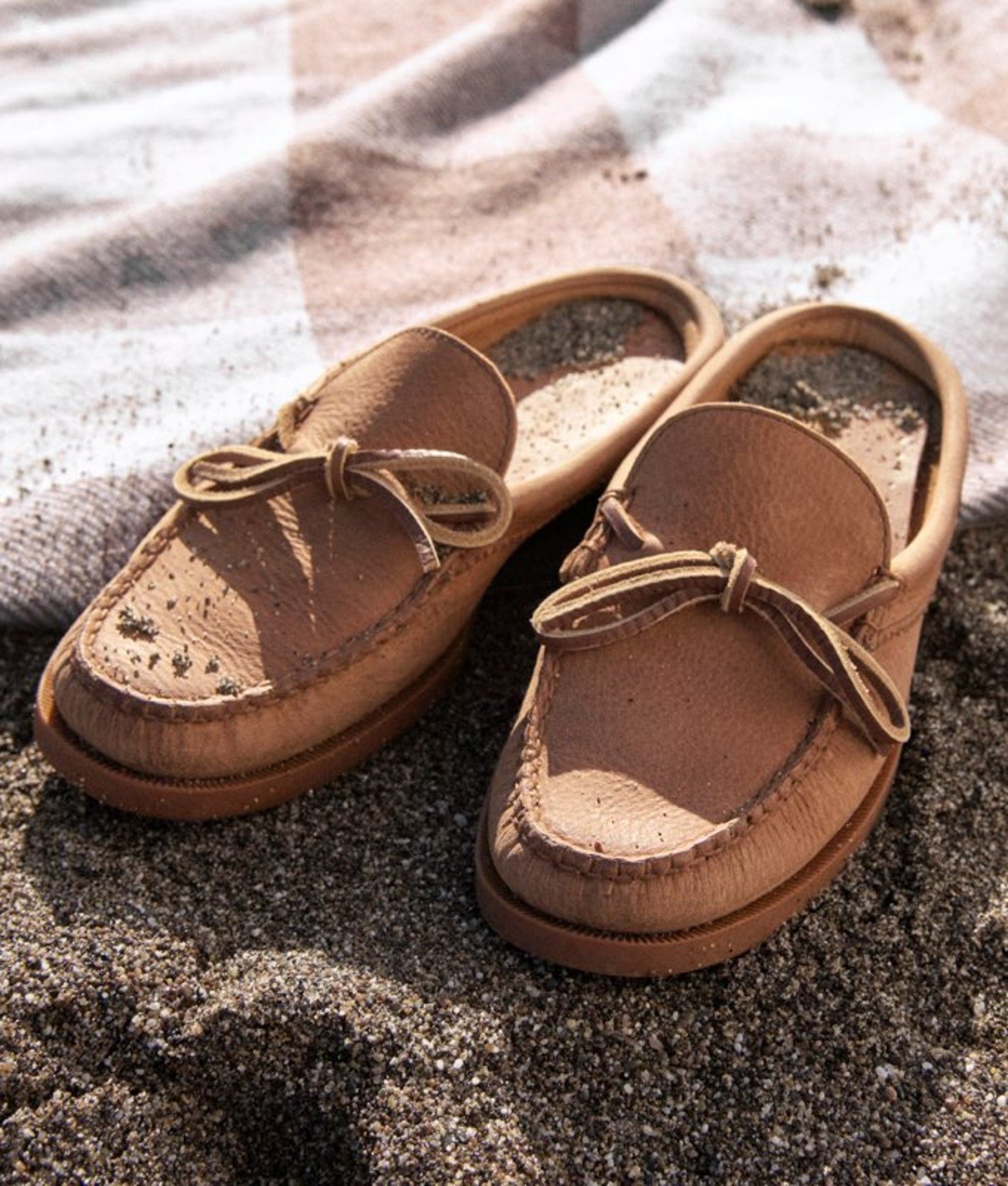 Pair of brown casual shoes placed on sand near the water’s edge. 